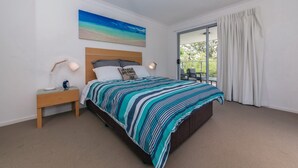 Quarto com cama, mesa de cabeceira, luminária, quadro e vista por uma porta de vidro.