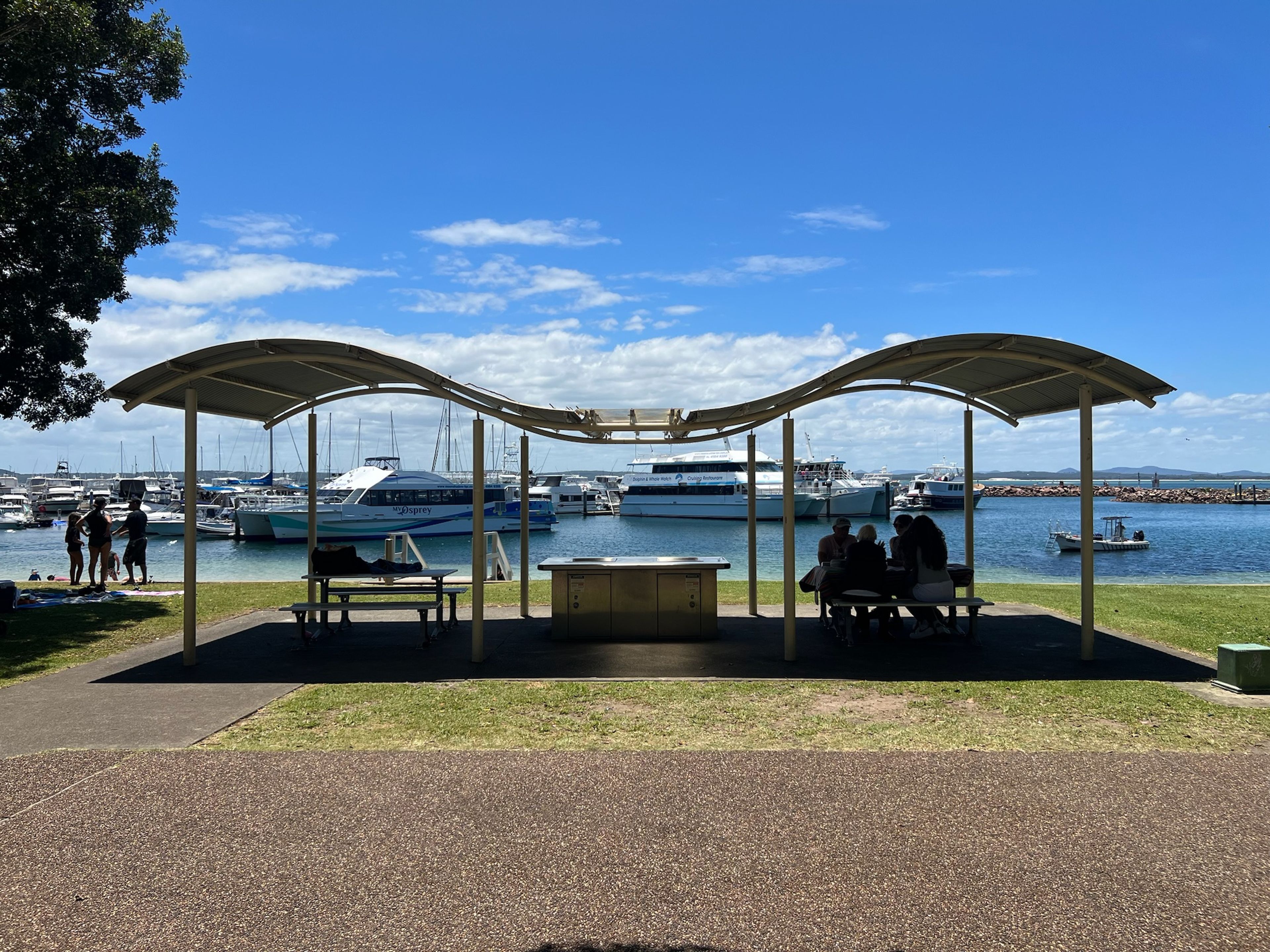 A covered outdoor seating area with tables and benches, overlooking a marina with boats.