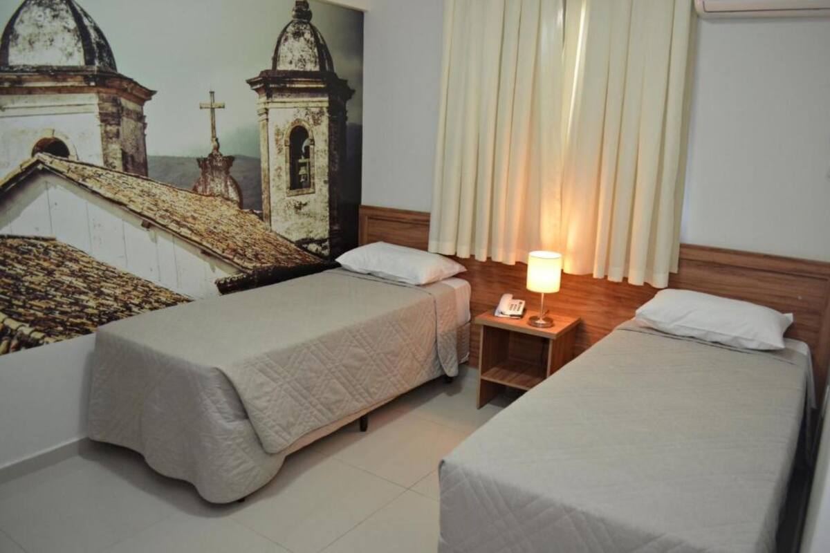 A hotel room with two single beds, a nightstand with a lamp, and a wall mural of a church.