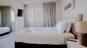 Habitación de hotel con una cama, un televisor montado en la pared, una ventana con cortinas y una mesita de noche con lámpara y control remoto.