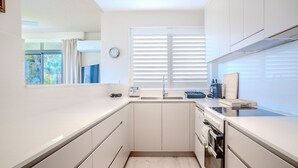 A compact kitchen with white cabinetry, a built-in oven, and a window with blinds. - The Helm, 1/22 Voyager Cl - Cross the grassy reserve and you're on beautiful Shoal Bay Beach! (Nelson Bay)