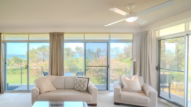A spacious living room with a large sliding glass door leading to a balcony, a ceiling fan, and a glass coffee table. - The Helm Waterfront Apartment in Nelson Bay (Nelson Bay)