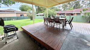 Een terras buiten met een houten tafel en stoelen, een barbecue en een overdekt gedeelte met uitzicht op een woonwijk.