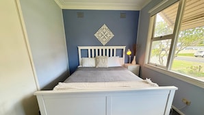 Een slaapkamer met een wit bed, een nachtkastje met een lamp en een raam met uitzicht op het exterieur.