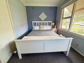 Quarto com cama branca, mesa de cabeceira com abajur e janela com vista para o exterior.