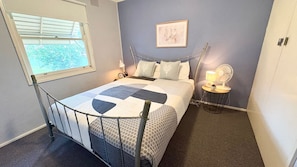 Une chambre à coucher avec un lit, une table de chevet avec une lampe, un tableau accroché au mur et une fenêtre avec des stores.