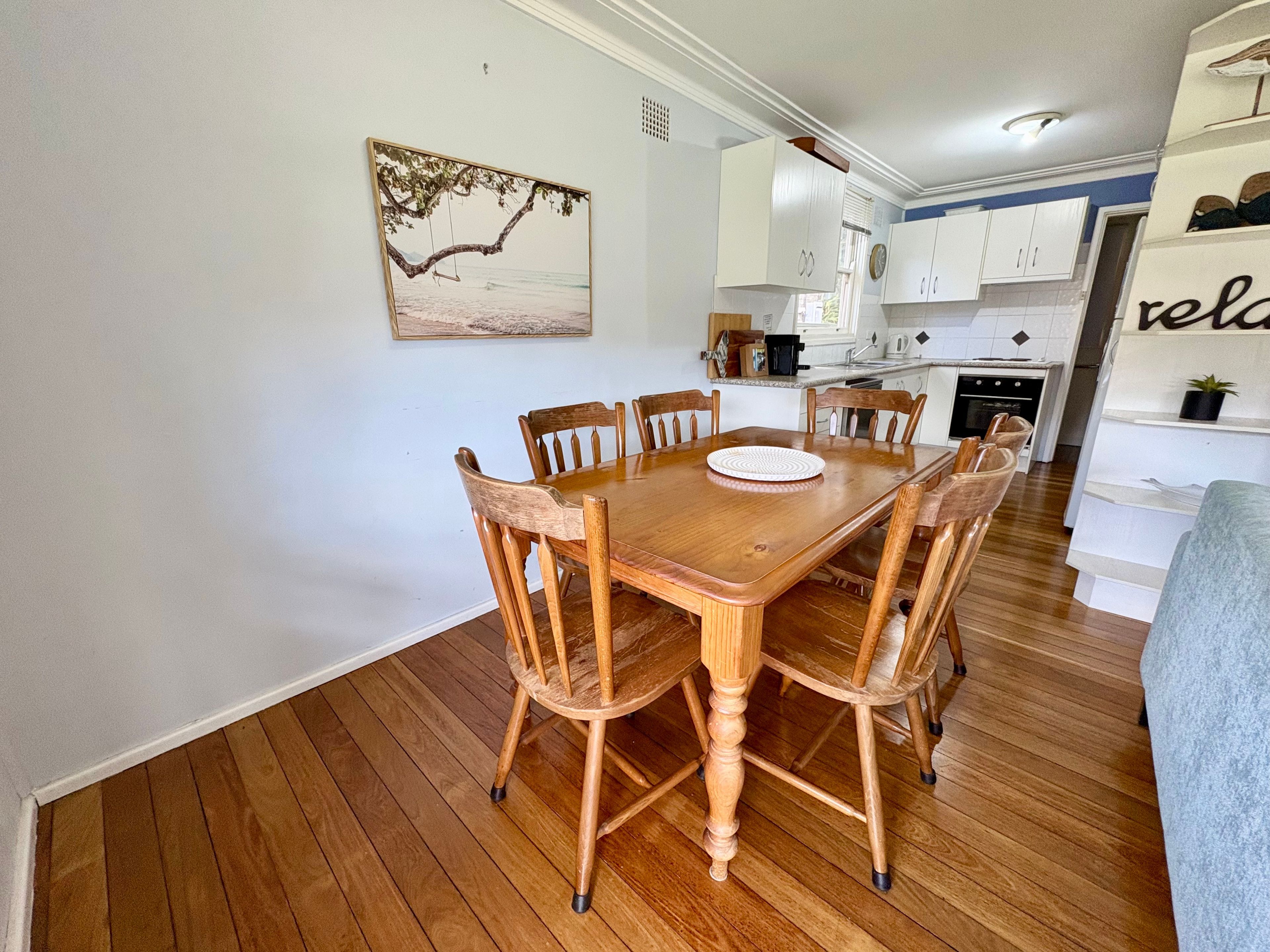 A dining area with a wooden table and chairs, a kitchen in the background, and a wall-mounted picture.