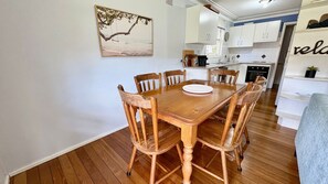 A dining area with a wooden table and chairs, a kitchen in the background, and a wall-mounted picture.