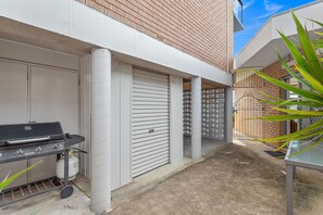 A patio area with a barbecue grill, a closed garage door, and a potted plant. - Bella Vista 22 Waterfront Apartment in Shoal Bay (Shoal Bay)