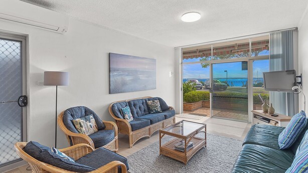 Un salon spacieux, meublé avec des meubles en osier, une porte vitrée donnant sur un balcon avec vue sur la mer, et un téléviseur à écran plat fixé au mur.