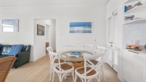 Un salon moderne avec une table ronde en verre entourée de chaises blanches, un canapé vert avec des coussins à rayures et un fauteuil en osier. Deux tableaux encadrés ornent le mur, ainsi qu’une porte blanche et un coin cuisine avec un meuble blanc et une étagère où sont disposés des objets décoratifs.
