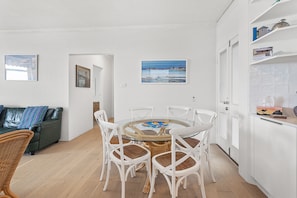 A modern living room with a round glass table surrounded by white chairs, a green sofa with patterned cushions, and a white cabinet with decorative items.
