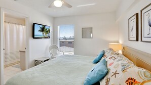 Un dormitorio con cama, ventilador de techo, televisor, ventana con persianas y cuadros en la pared.
