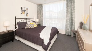 Una habitación de hotel con cama, mesitas de noche, lámpara, ventana con cortinas y una obra de arte colgada en la pared.