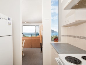 A compact kitchen with a stove, a window with a view of the sea, and a small potted plant on the windowsill. - Ocean Shores Stunning Waterfront Nelson Bay Escape (Nelson Bay)
