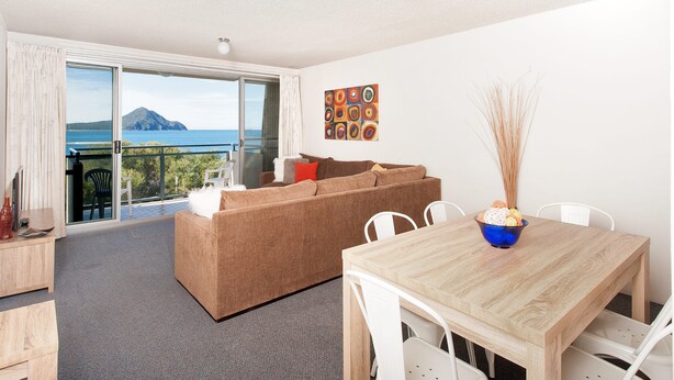 Una sala de estar moderna con sofá, mesa de comedor y sillas, un balcón con vista al mar y un cuadro en la pared.