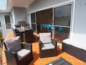 Een terras buiten met rieten meubilair, een barbecue en een schuifpui naar een eethoek binnen.