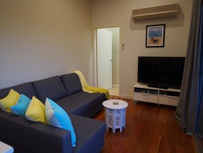 A living room with a sofa, a television, a small white table, and a door leading to another room. - Pollys' View Nelson Bay Water View Family Home (Nelson Bay)