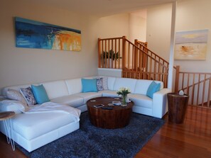 A modern living room with a white sectional sofa, a wooden coffee table, and a staircase with wooden railings. - Pollys' View Nelson Bay Water View Family Home (Nelson Bay)