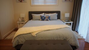 A neatly arranged bedroom with a large bed, two bedside tables, a bedside lamp, a window with blinds, and a curtain.