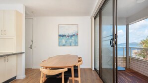 Uma varanda com mesa e cadeiras de madeira, porta de vidro que dá para um terraço e um quadro na parede.