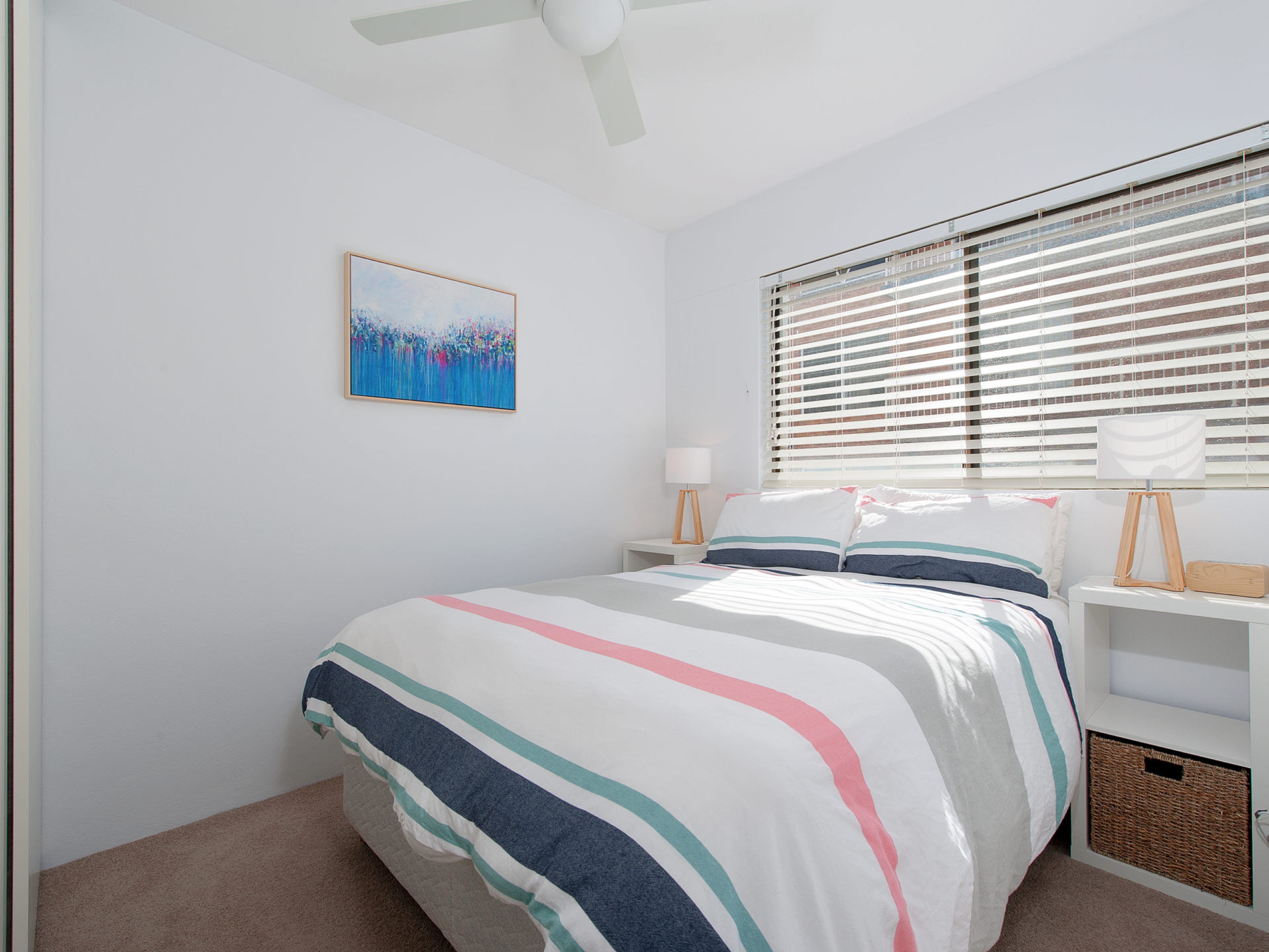 A bedroom with a bed, a nightstand, a lamp, a picture on the wall, and a window with blinds.