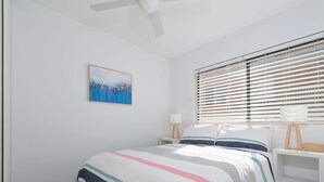 Quarto com cama, mesa de cabeceira, abajur, quadro na parede e janela com persianas.