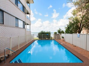 A rectangular swimming pool with a metal safety fence, a brick building with windows, and a clear sky. - Bangalee 15 with Pool and Beach Access (Soldiers Point)