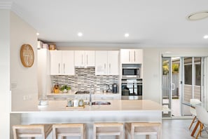 A modern kitchen with white cabinetry, a central island, and a dining area with a glass door leading outside. - Signature House Family Retreat Corlette (Corlette)