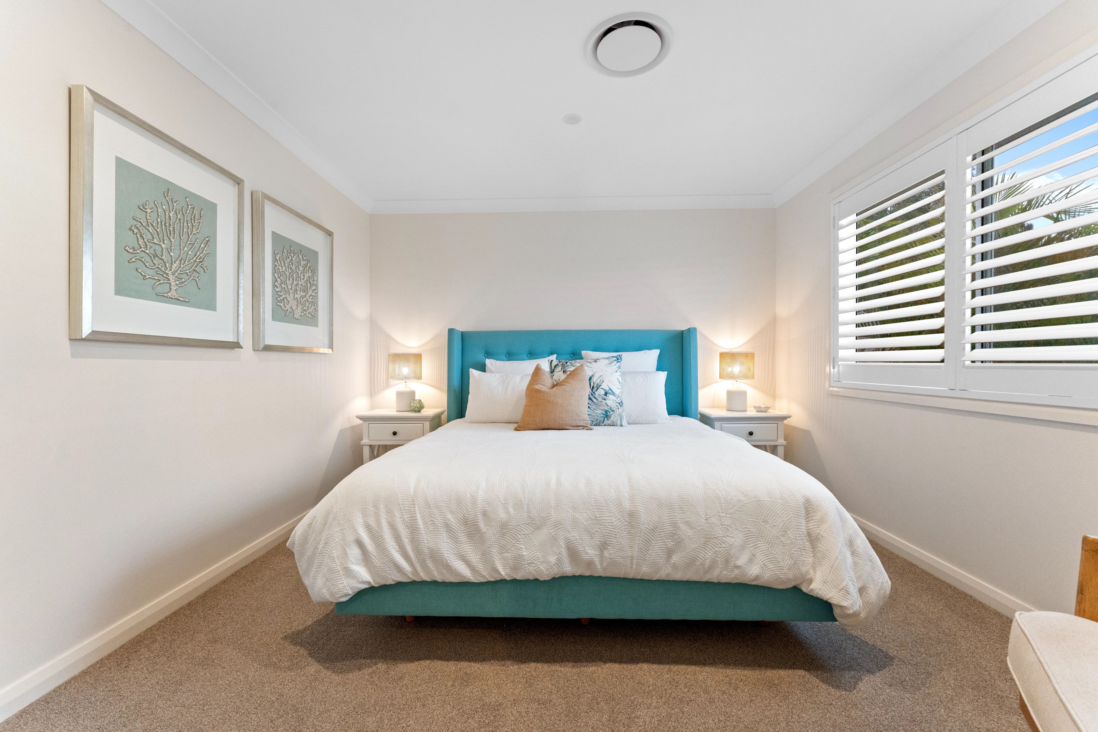 A bedroom with a bed, two bedside tables, a window with blinds, and two framed pictures on the wall.