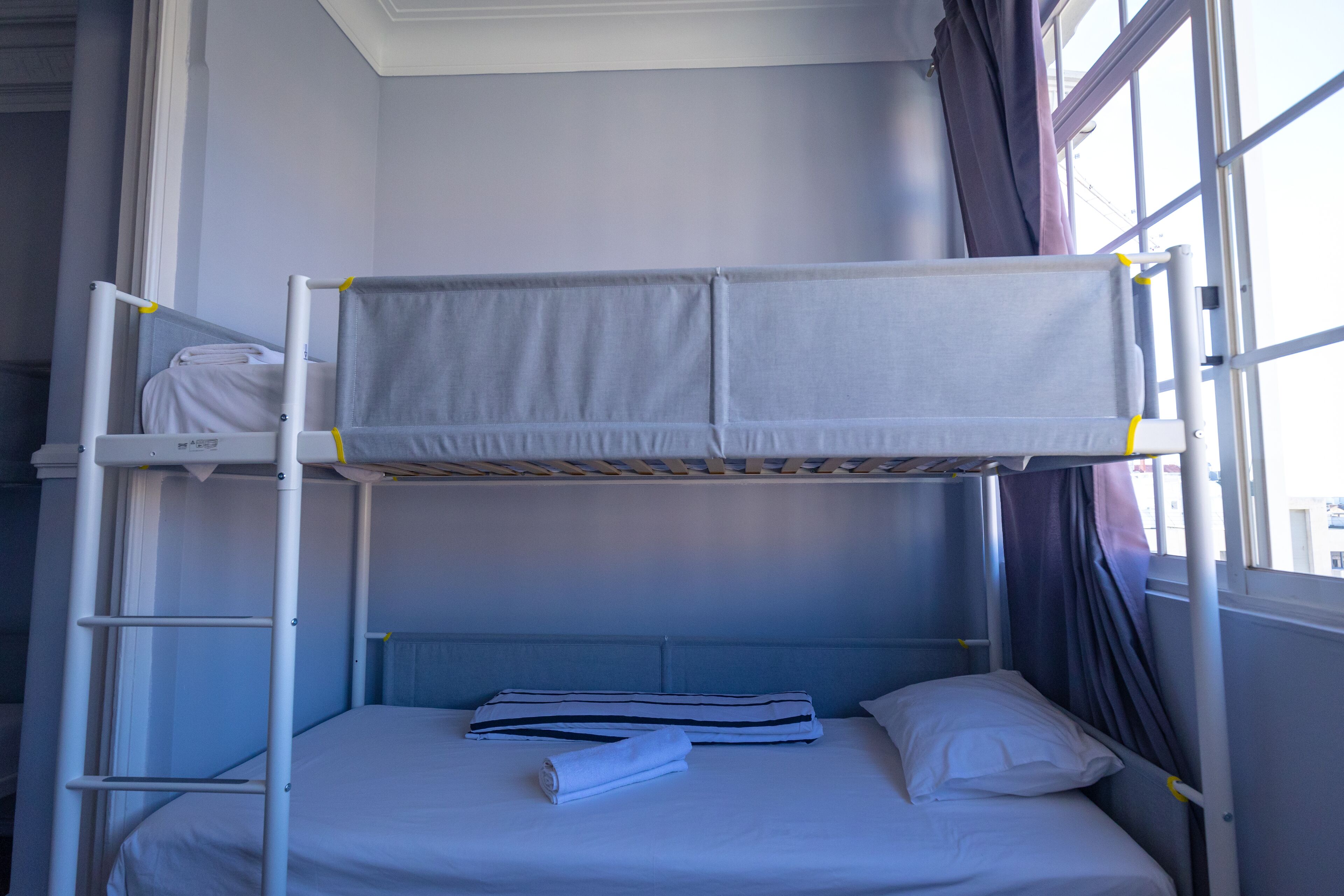 Une chambre avec un lit superposé, une échelle, une fenêtre avec des rideaux et un lit bien fait, avec des oreillers et une serviette.