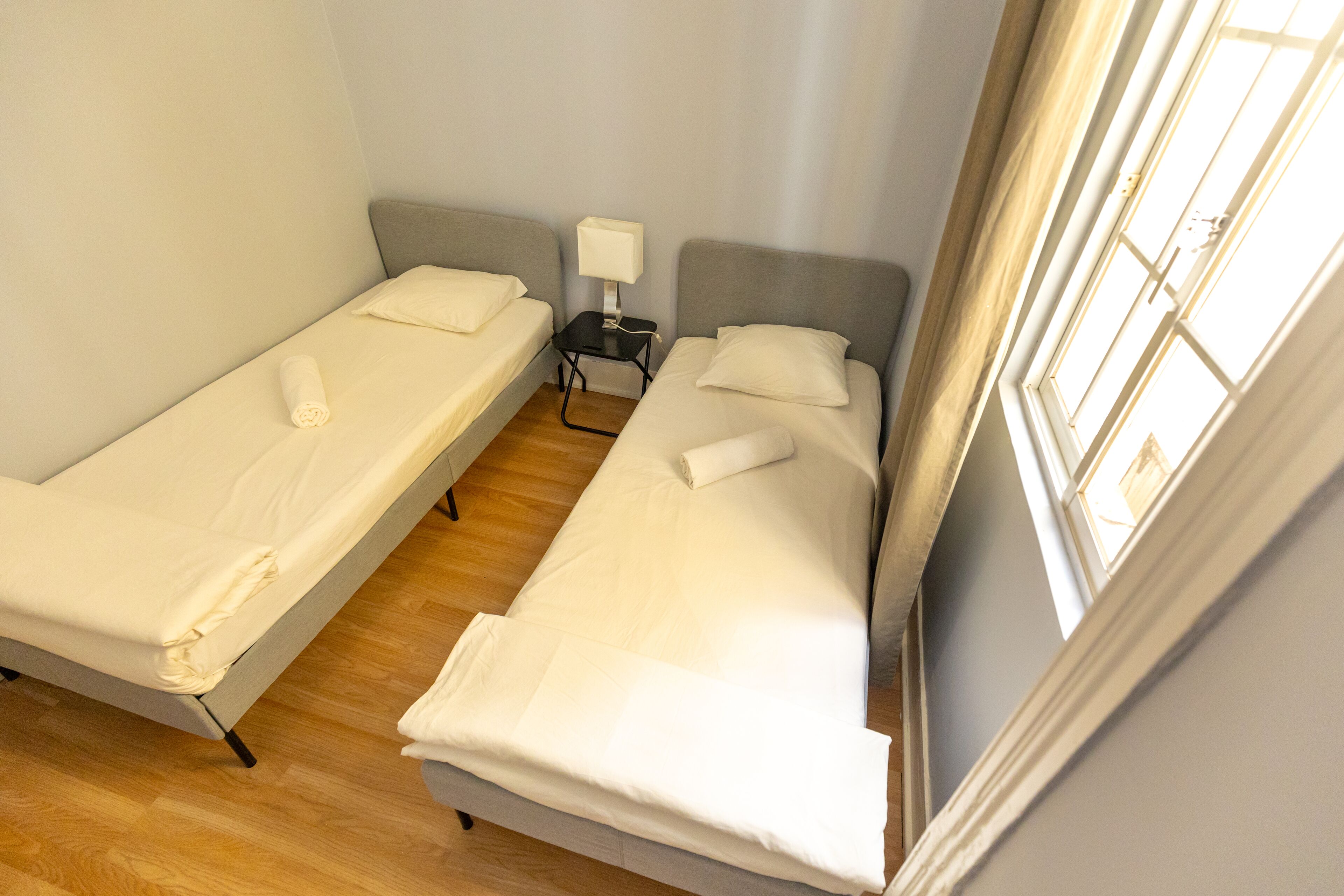 A room with two single beds, a small table with a lamp, and a window providing natural light.