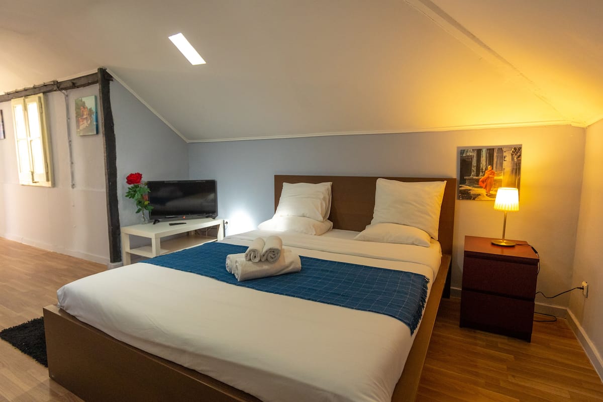 Una cama bien hecha con una manta azul, un televisor sobre una mesita y una lámpara en una mesita de noche.