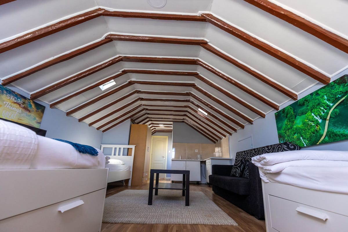 Habitación compacta con techo inclinado, una cama, una mesita, un sofá y un televisor.