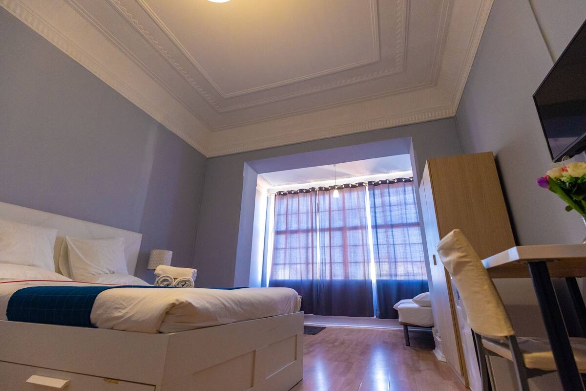 Habitación de hotel con cama, escritorio, silla, televisor y ventana con cortinas.