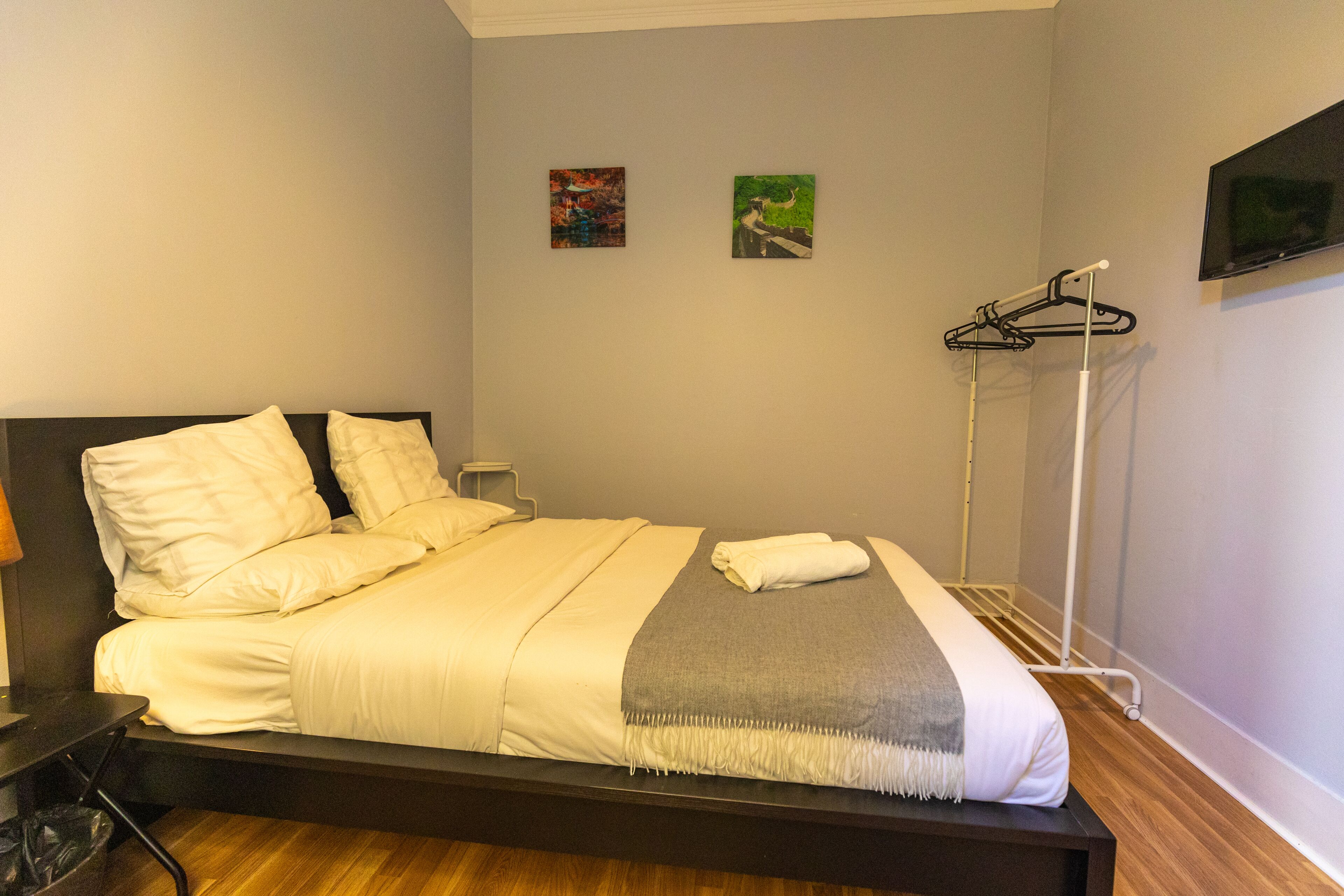 A neatly made bed with a grey blanket and white pillows, a bedside table with a lamp, and a wall-mounted TV.