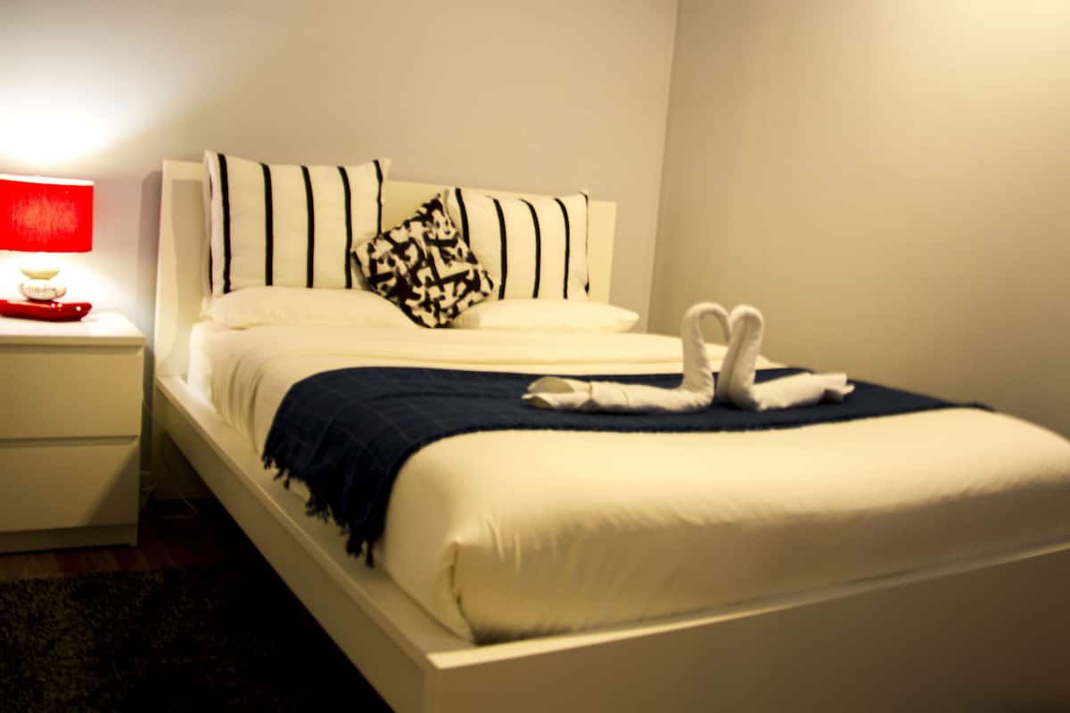 Una cama bien hecha con ropa de cama blanca y una manta azul, un cojín con diseño blanco y negro y una toalla con forma de cisne sobre la cama.