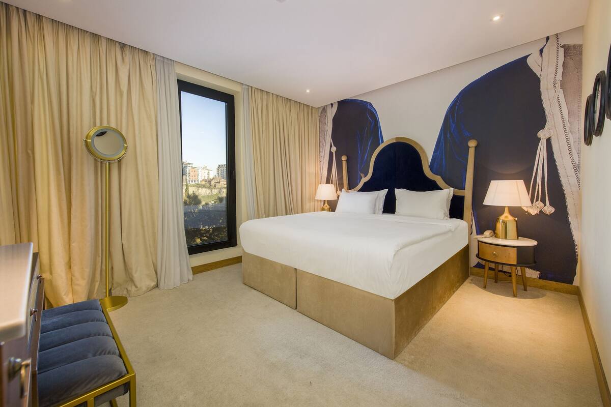 Une chambre d’hôtel avec un grand lit, une table de chevet avec une lampe, une chaise et une vue sur la ville par une grande fenêtre.