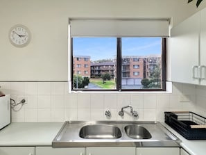 A kitchen with a double sink, a window with a view of buildings, and a wall-mounted clock. - Foreshore Paradise Nelson Bay (Nelson Bay)