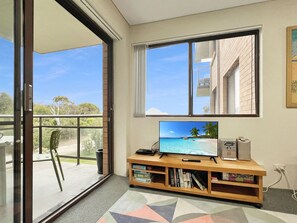 A room with a large window, a flat-screen TV, a bookshelf, and a view of a beach.