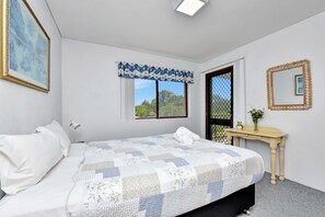 Een slaapkamer met een bed, nachtkastje, spiegel en raam met uitzicht op groen.
