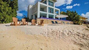 Una moderna propiedad frente al mar de dos plantas con un muro de contención de piedra, múltiples sillas de descanso y una vista despejada del océano.