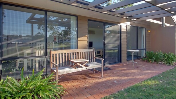 A modern building with large glass windows, a wooden bench, and a small table on a brick patio. - Horizons Retreat (Salamander Bay)