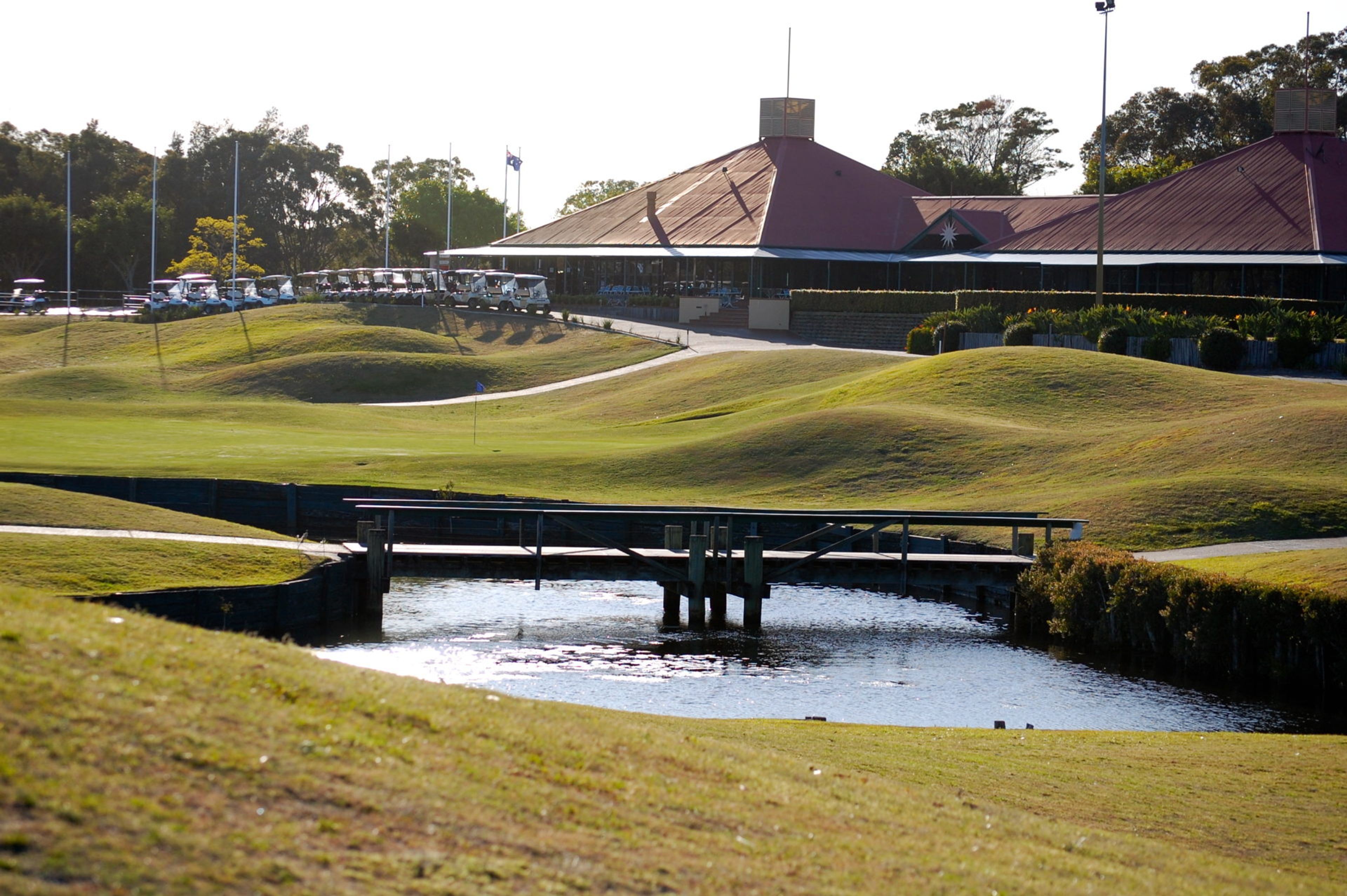 Un campo de golf con un puente sobre un canal de agua, rodeado de césped cuidado y colinas.