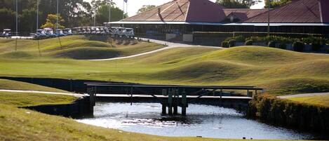 Un campo de golf con un puente sobre un canal de agua, rodeado de césped cuidado y colinas.