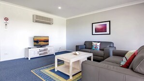 A living room with a sofa, a coffee table, a TV on a white cabinet, a painting on the wall, and a no-smoking sign. - Horizons Retreat (Salamander Bay)