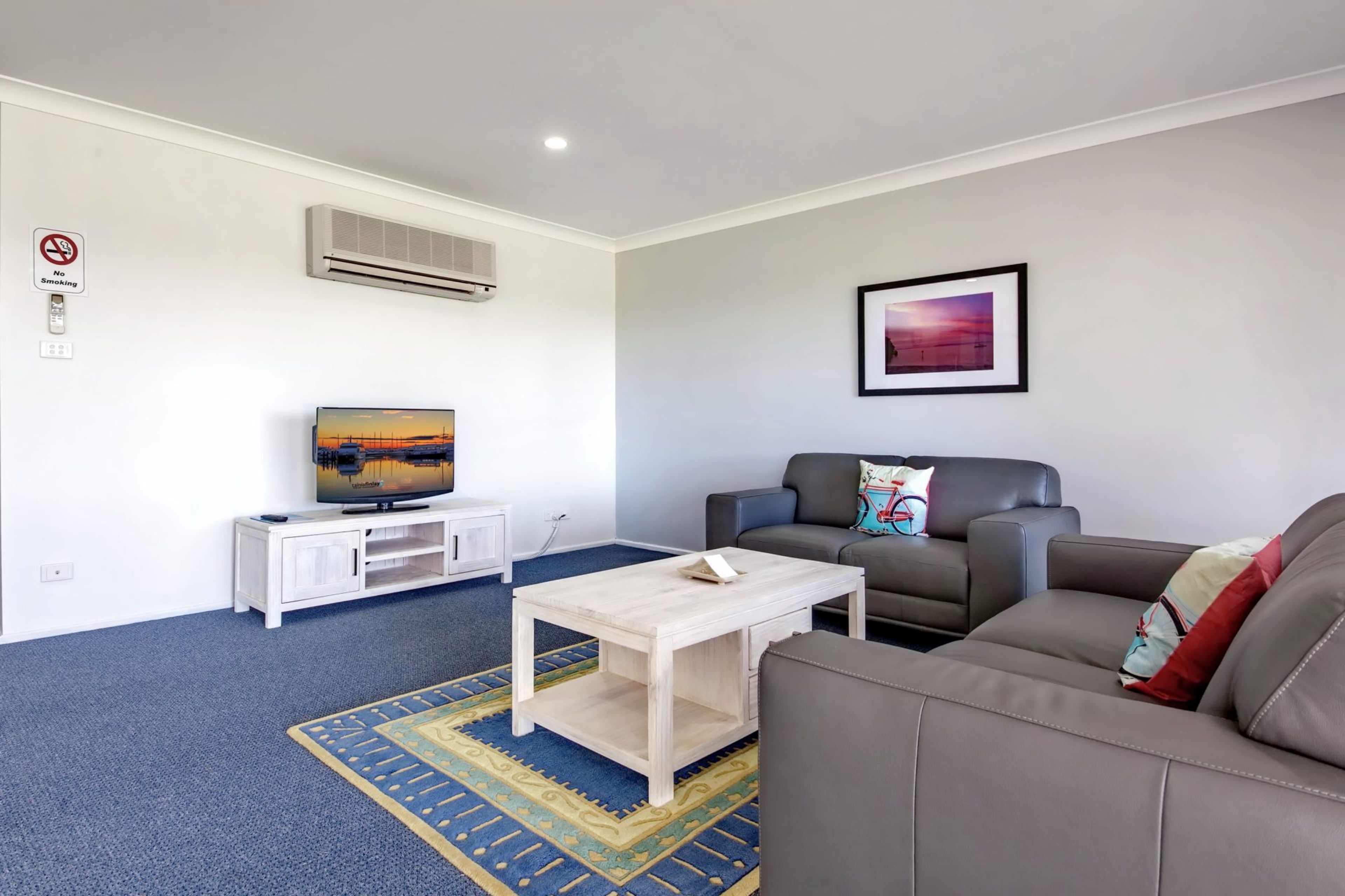 A modern living room with a flat-screen TV on a white stand, a grey sofa, a coffee table, and a patterned rug.