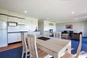 A modern kitchen with white cabinets, a dining table with chairs, a living area with a sofa and a TV, and a blue carpet.