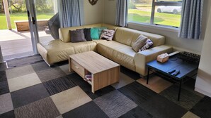 Un salon avec un canapé d’angle beige, une table basse en bois, une petite table noire avec une télécommande, et une vue sur un espace verdoyant grâce à de grandes fenêtres.