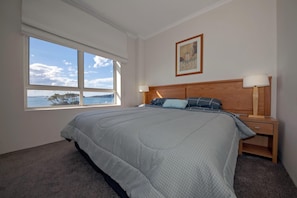 A bedroom with a large bed, a nightstand, a lamp, a window with a view of the sea, and a framed picture on the wall.